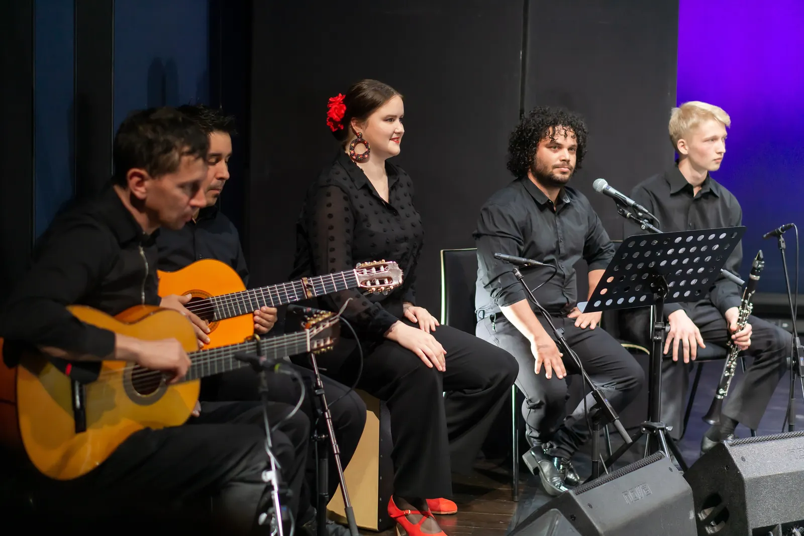 Two guitarists, a seated singer with a red flower in her hair, a second singer and a clarinettist on a dark stage